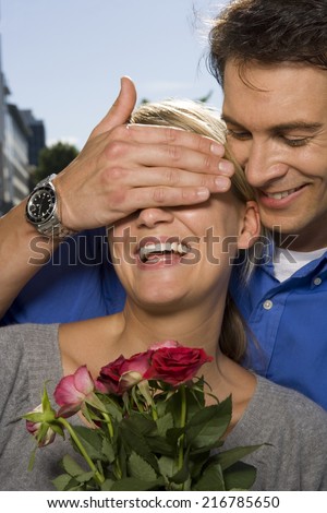 A man covering a woman\'s eyes and giving her roses.