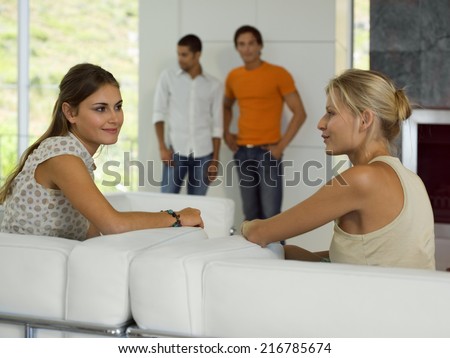 Women talking on the sofa, Men standing in the background.