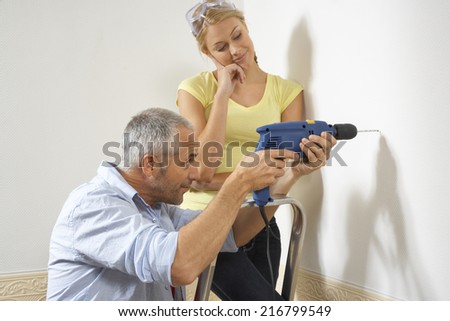 Side profile of a mid adult man drilling into a wall with a young woman standing beside him