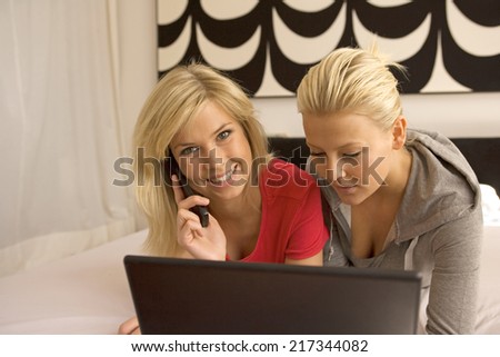 Portrait of a young woman lying in front of a laptop with another young woman talking on a flip phone