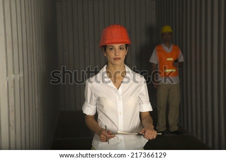 Portrait of a female dock worker holding a clipboard and a man standing behind her in a cargo container