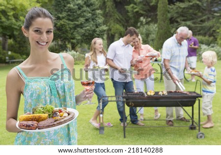 Portrait of smiling woman holding plate of barbecue and glass of wine with family in background