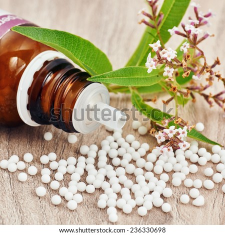 Homeopathic granules scattered around a glass bottle and a medicinal plant on a wooden table. Vertical image with empty space in the bottom for written messages and titles