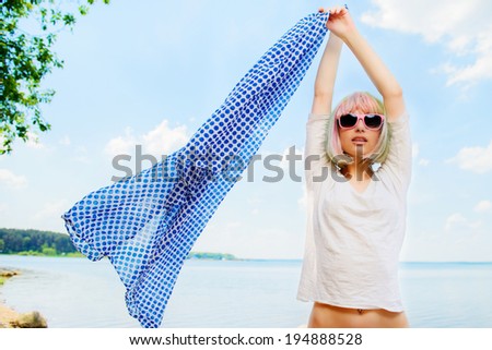 Portrait of beautiful sexy girl in a blue shirt and pink glasses on a background of blue sky and sea with his arms raised up holding a blue cloth