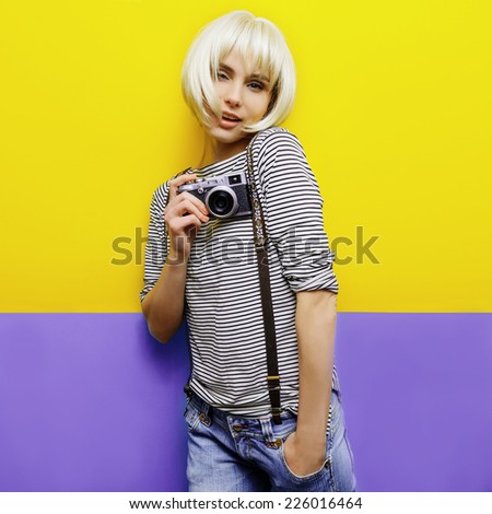 isolated studio portrait of young blond woman on yellow-purple background wearing jeans, a striped shirt and suspenders holding retro camera in hand