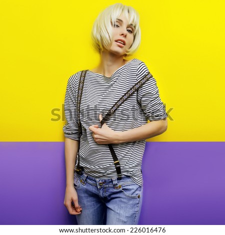 isolated studio portrait of young blond woman on yellow-purple background wearing jeans, a striped shirt and suspenders