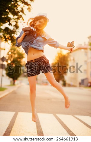 portrait of a beautiful positive jumping girl in  hat holding a camera in his hands on a background of the sunny city