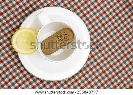 a tea cup with a cookie in it and a piece of lemon at the checked cloth background