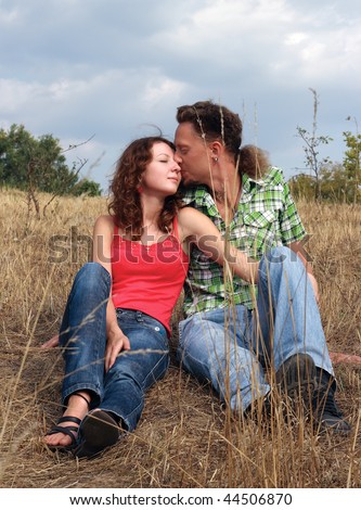 Beautiful young woman with curly hair and man sit in the field. The young man is kissing the lady on her cheek.