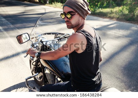 Handsome man biker posing outdoor. Sitting on bike and wearing stylish outfit and mirrored sunglasses.