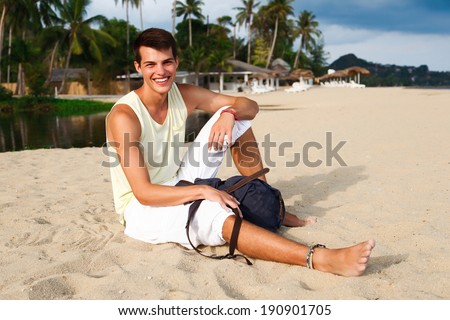 Outdoor fashion portrait of young handsome man posing at beach in tropical country with his backpack.