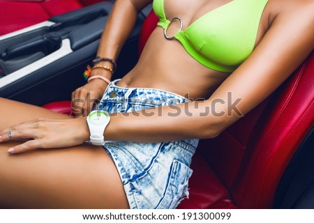 Close up image of young sexy woman sitting in the car in bright bikini and high waisted jeans shorts.