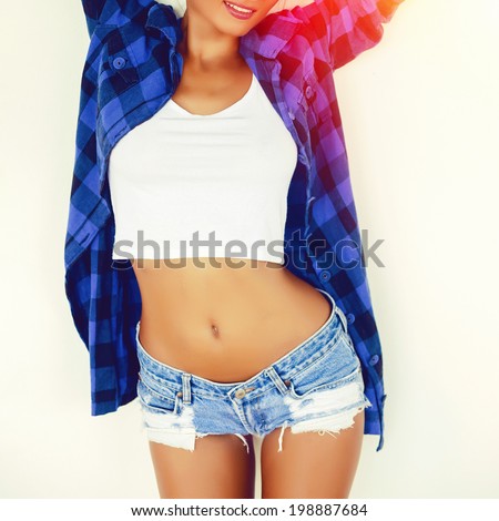Close up fashion image of young sexy tan sportive woman posing in stylish jeans shorts near white wall. Bright instagram colors.