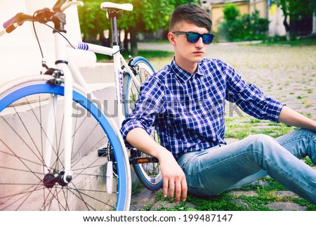 Young stylish hipster man sitting on the grass in old european city park with his bike, wearing stylish retro outfit and sunglasses.