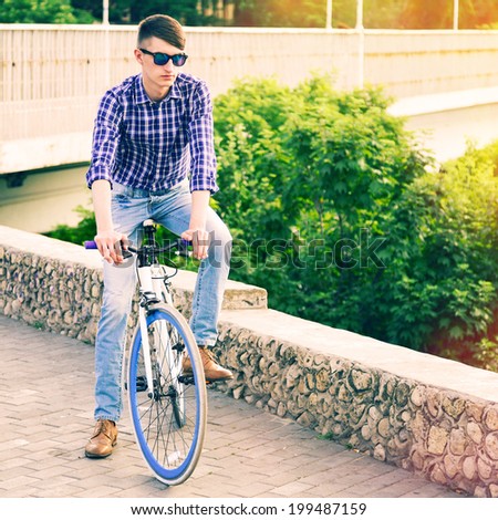 Young handsome man posing outdoor in summer time near green park at sun sine, riding stylish blue modern fixed bike.