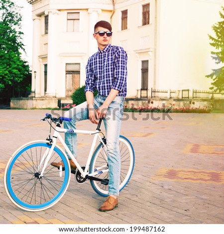 Bright portrait of young handsome stylish hipster man posing on blue fix bike at old european city. Warm sunshine colors.