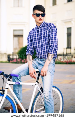 Young handsome man posing city on his stylish bike, wearing navy blue sunglasses and plaid shirt.