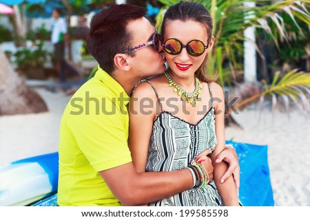 Handsome man kissing to the cheek pretty young brunette, wearing summer bright stylish clothes and mirrored sunglasses, having fun together at romantic trip.