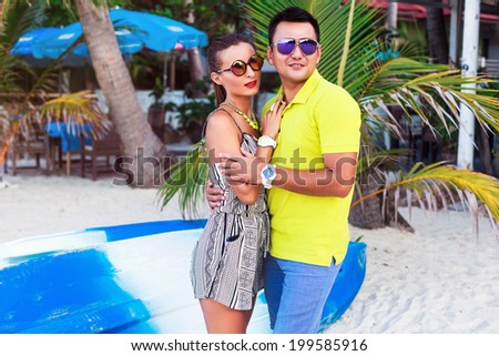Summer portrait of happy couple in love posing near palms and colorful kayak boats, in stylish bright neon clothes and mirrored sunglasses.