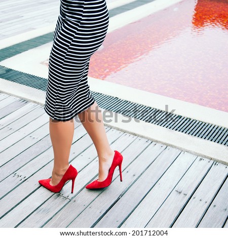 Woman in stripy black and white dress posing near creative red pool in height heeled classic pumps. Fashion image.