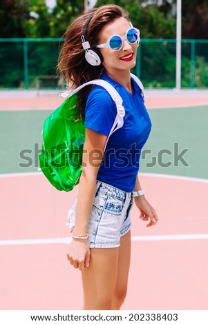 Young pretty brunette woman jumping in american sports ground, wearing bright clothes, mirrored blue sunglasses,neon clear backpack and white earphones, listening music and having nice day.