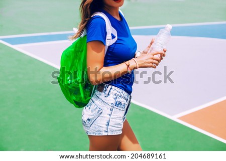 Young slim tan woman, posing alone at sports ground after had fitness training in stylish bright summer casual outfit and big bottle of mineral water.