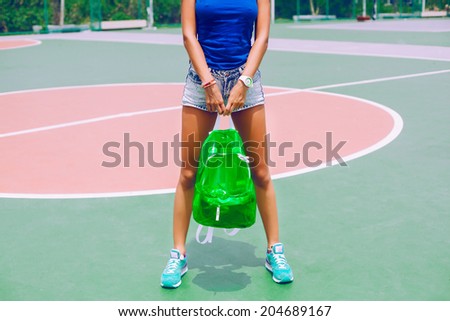 Outdoor image of woman with long tan sportive legs, wearing short vintage jeans shorts, holding her clear neon backpack and posing at american sports ground.