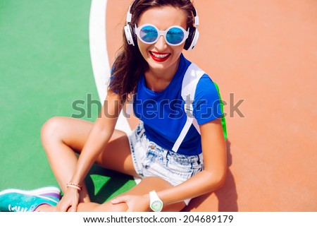 Outdoor fashion portrait of hipster girl posing at sports ground in bright summer outfit, listening music and wearing stylish sportive shoes backpack and sunglasses.