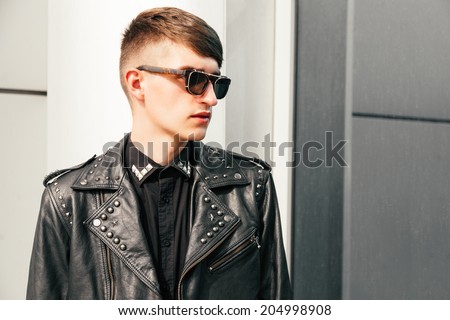 Handsome stylish guy in trendy black leather jacket designer sunglasses and hipster haircut posing outdoor at urban background.