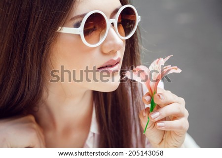 Young pretty girl posing in summer sunny day with small beautiful flower, have natural melt up and manicure,brunette straight hair and round sunglasses.Soft tender colors.