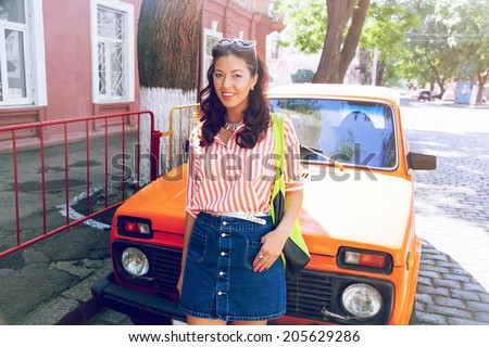 Happy brunette woman with retro curl hair style posing at the street near big old orange retro car, enjoy summer hot day and her summer holidays.
