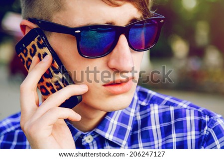 Close up fashion portrait of stylish handsome hipster guy speaking with friends on his smart phone, wearing blue plaid shirt and mirrored sunglasses.