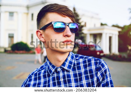 Outdoor fashion portrait of young handsome hipster man with stylish haircut posing at city in plaid shirt and mirrored sunglasses.