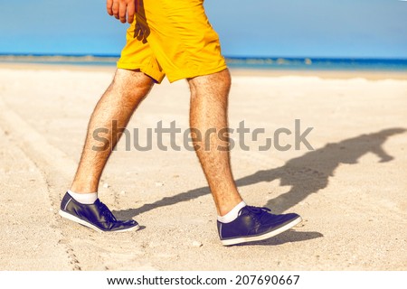 Close up fashion image of man walking alone at tropical exotic beach with blue ocean and white sand, wearing stylish shorts and sneakers.