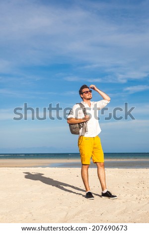 Beach guy enjoying sun tanning on travel smiling under blue sky. Handsome man having fun on tropical beach. Wearing backpack yellow shorts and white shorts.