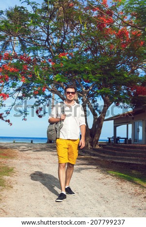 Outdoor fashion portrait of man traveling at ho exotic country. Beautiful view on ocean beach and tropical plants. Wearing bright casual outfit and sunglasses.