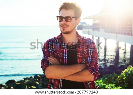 Handsome stylish man in hipster plaid shirt and sunglasses posing near sea side at sunshine, enjoying beautiful view and relax near ocean.