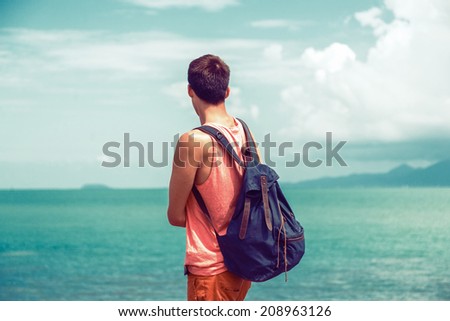 Young traveler man with backpack staying alone at seaside and looking to the ocean, exploring the world fell new emotions, vintage instagram colors, deep blue sky and sea water.