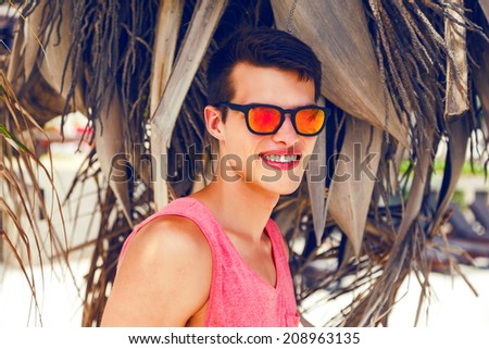 Close up fashion portrait of handsome stylish guy posing at hot tropical exotic country near palm tree wearing mirrored sunglasses, have big pretty smile.