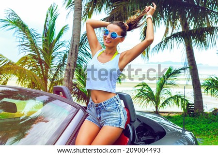 Pretty woman have fun at luxury car in vacation, posing near sea side with palm trees, wearing hight waisted shorts and stylish round sunglasses.