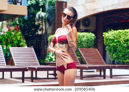 Fashion portrait of sexy woman with perfect tan body posing in bikini at luxury resort, just coming from the pool with wet body and hairs. Retro sunglasses.