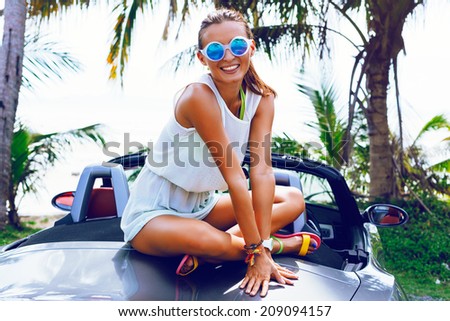 Pretty smiling girl sitting on luxury car, and having fun on vacation, wearing bright summery dress and sunglasses, palm trees background.