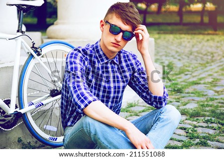 Close up fashion portrait of stylish handsome hipster guy sitting at city park near his fixed bike, wearing blue plaid shirt and mirrored sunglasses.