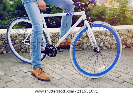Close up outdoor image of young hipster man in jeans and vintage shoes sitting at stylish modern fix bike. Sunny summer bright colors.