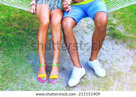 Close up fashion image of couple woman and man legs sitting at the beach, have perfect bronze tan, bearing casual white sneakers and bright neon flat sandals.
