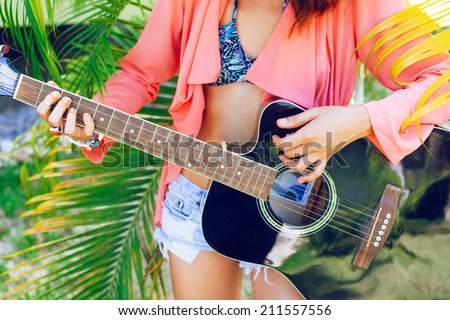 Close up fashion image of woman holding big black acoustic guitar, wearing vintage denim shorts, bright shirt bikini and accessorizes. Nice sunny hot day, palms garden location.