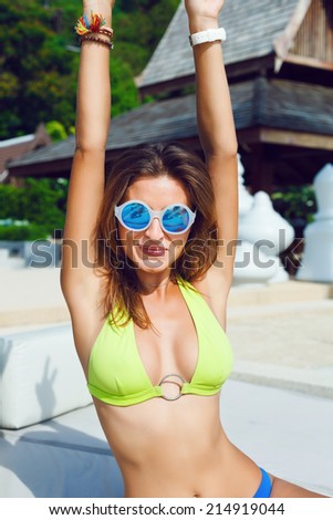 Happy young sexy tan fit woman relaxing and having fun at luxury beach resort, put her hands in the air and smiling. Wearing bright bikini and sunglasses.