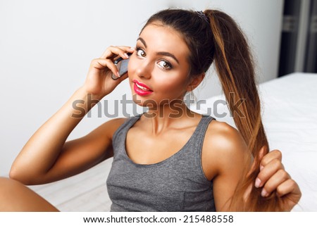 Young pretty woman with bright make up and perfect bronze skin speaking on smart phone, and holding her ponytail. Lifestyle indoor portrait.