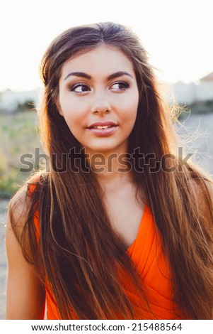 Fashion outdoor portrait of pretty woman wit amazing long straight brunette hairs big brown eyes and full sensual lips posing at countryside on sunset, wearing bright orange dress.
