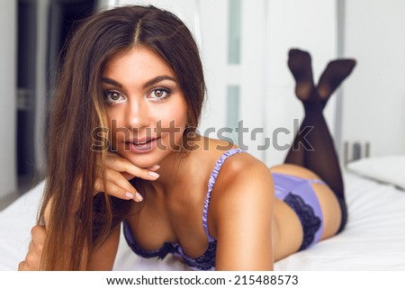 Indoor fashion portrait of amazing sensual beautiful woman with perfect glow skin natural make up and long brunette hairs, laying on be a at sexy silk luxury lingerie.